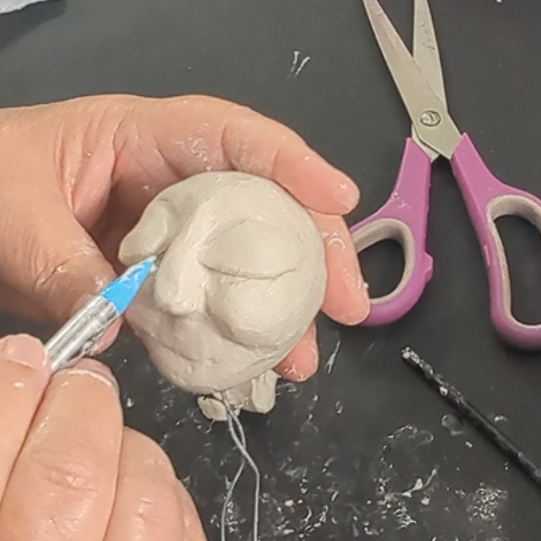 Sculpting the eye area of a handmade clay face with a small modeling tool.