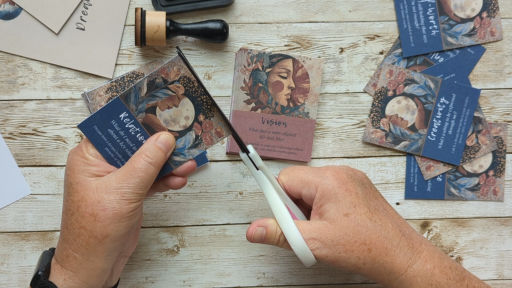 Hands cutting intuitive Dreamory Cards on a wooden table, surrounded by ink tools and finished cards for a soulful creative project.