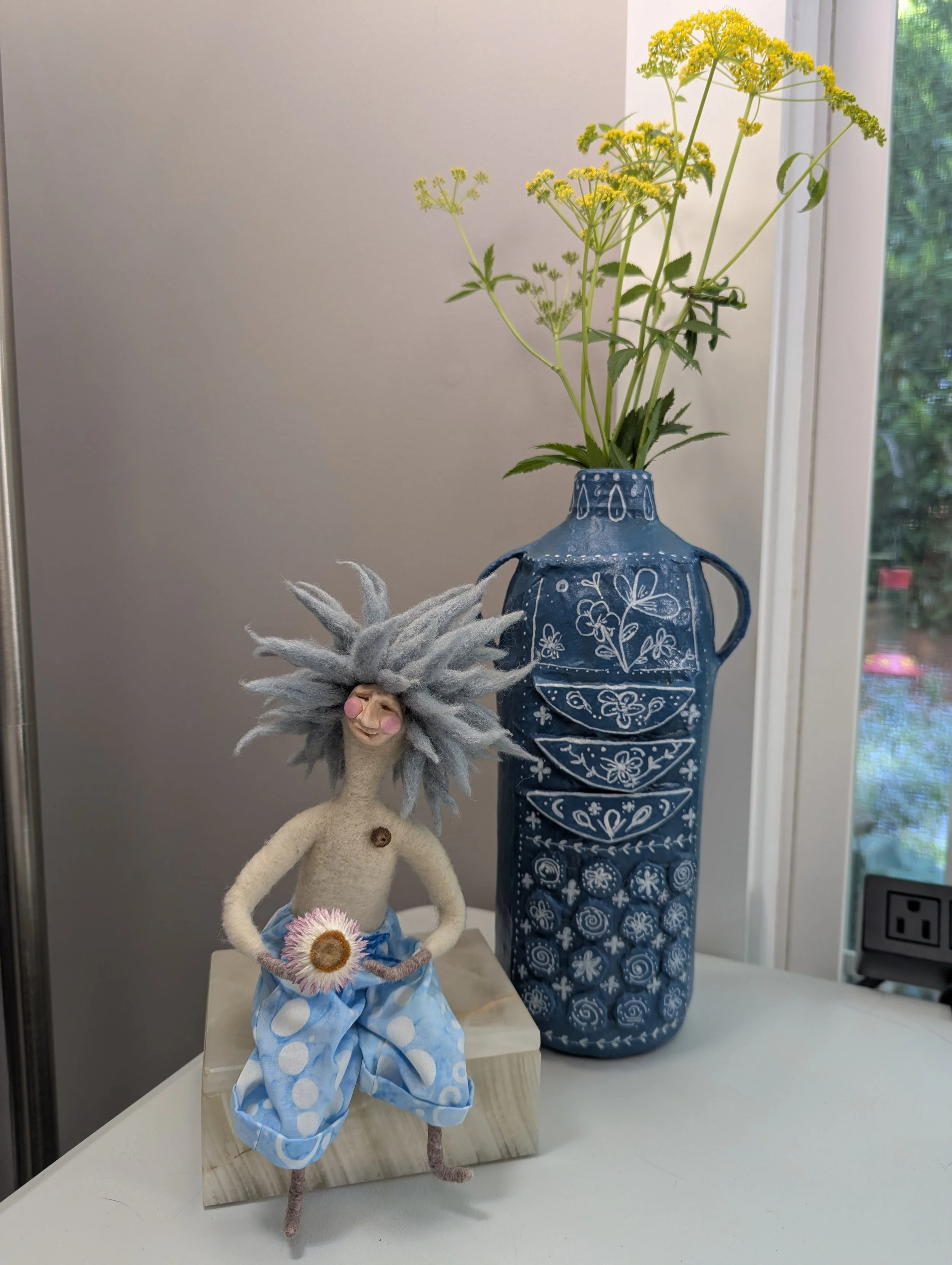 a blue papier mache vase with yellow flowers in it sitting by a window with a small needle felted doll with wild blue hair holding a flower