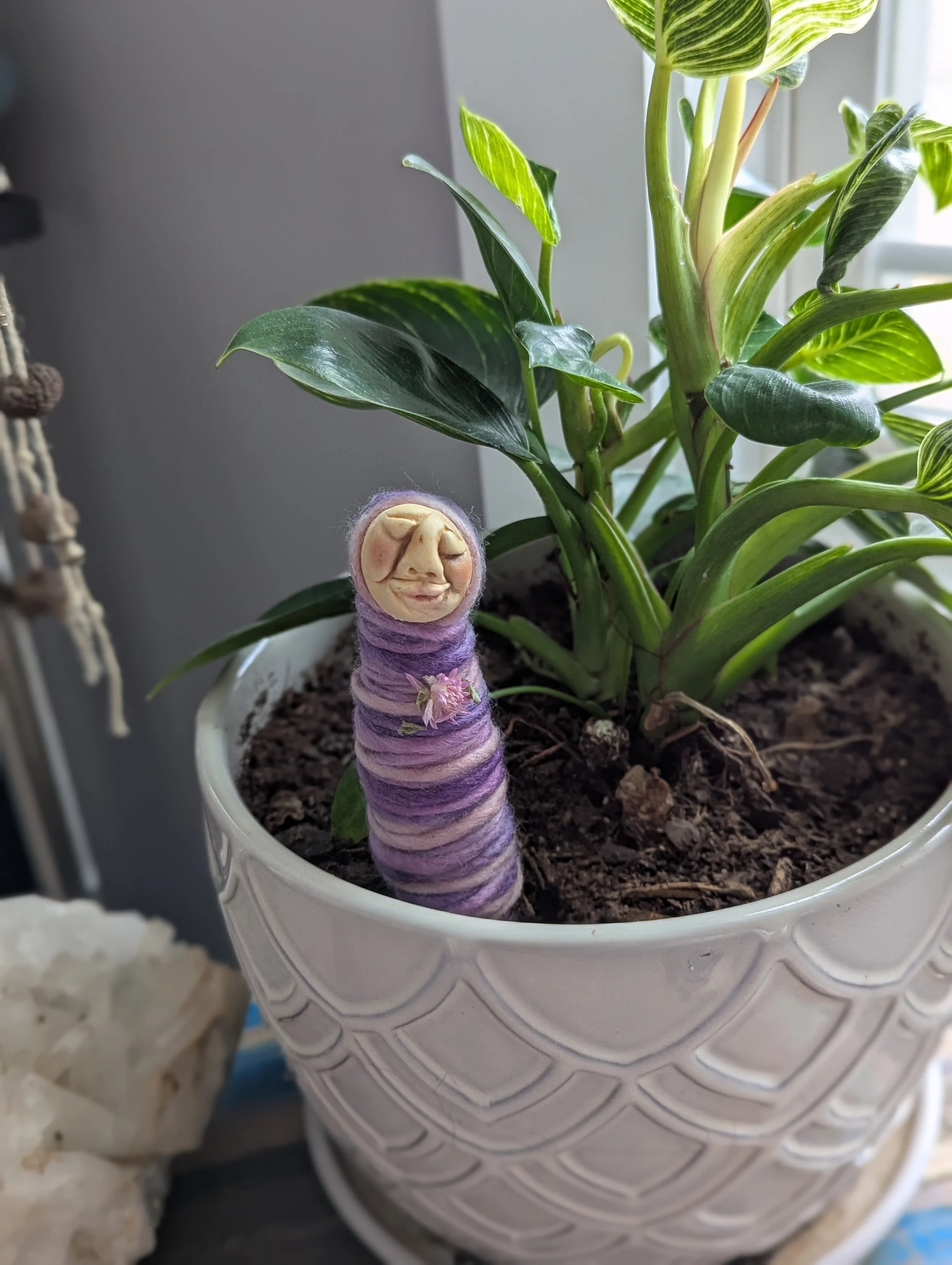 Plant person with an abstract clay face and multicolored yarn wrapping, standing in a potted plant with trailing greenery
