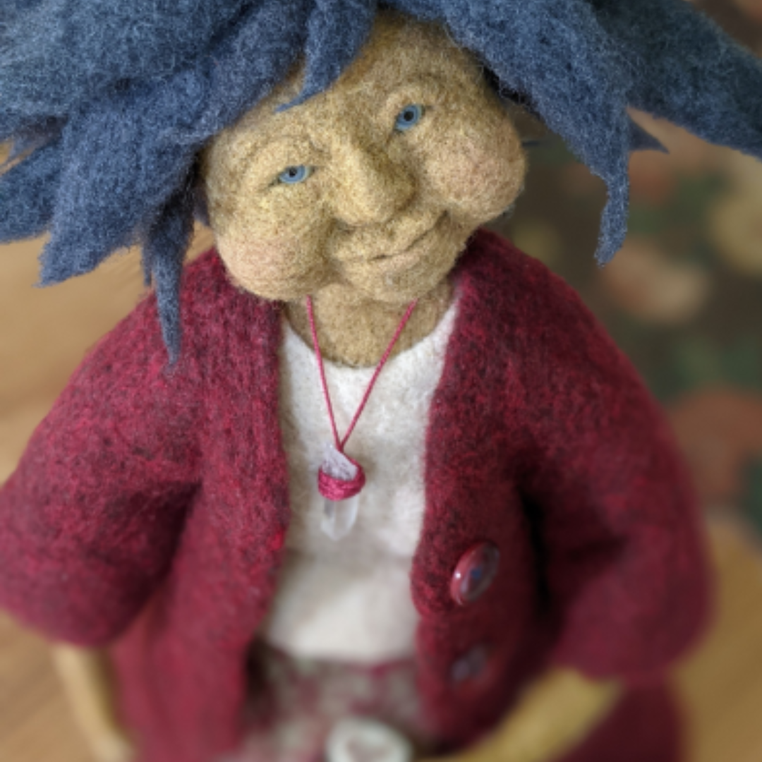 A close-up of a handmade spirit doll with soft felted features, blue wool hair, and a gentle smiling expression. The doll wears a deep red felted jacket, a white shirt, and a small crystal pendant on a pink cord.