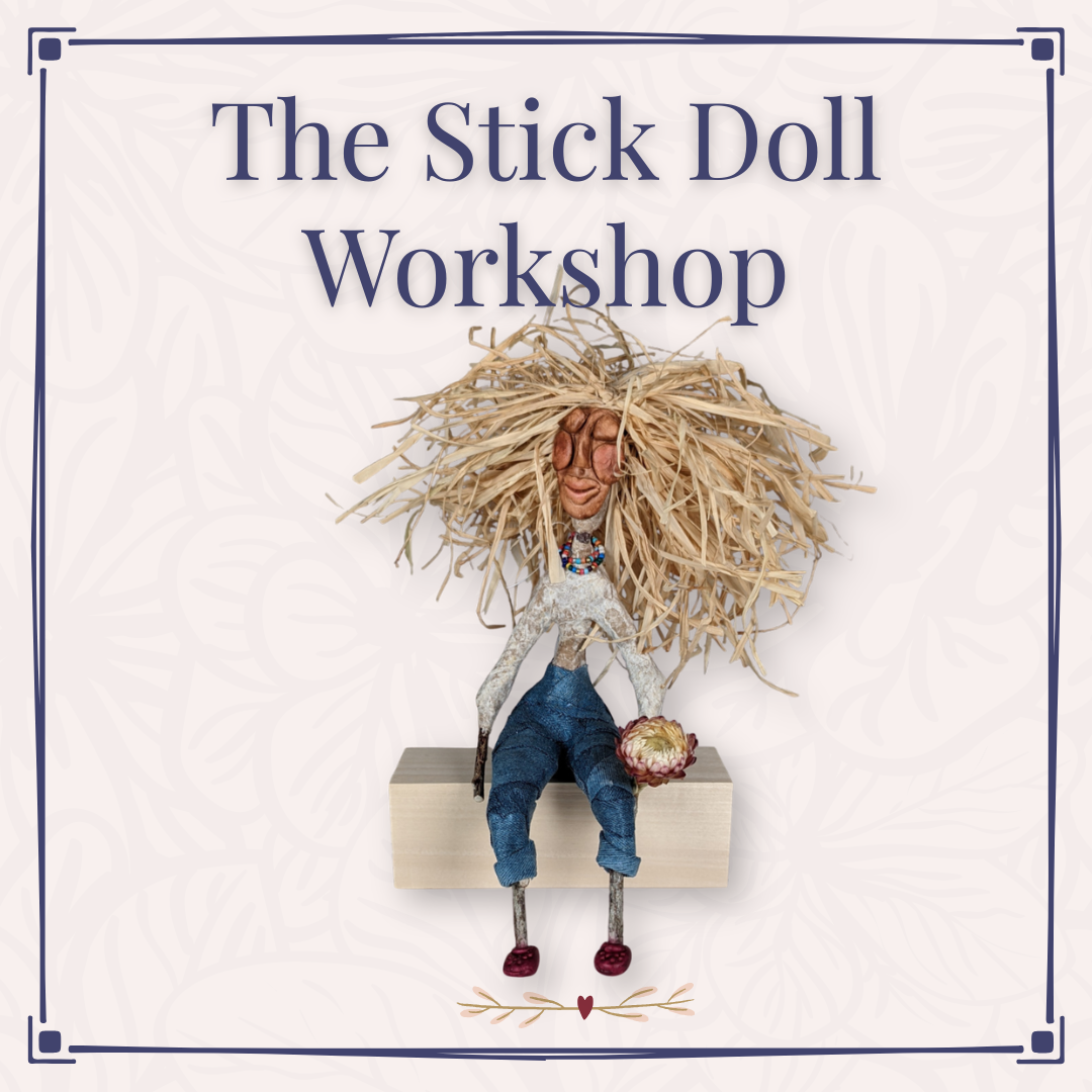 Handmade stick doll withblue pants, raffia hair sitting on a block of wood holding a dried flower.
