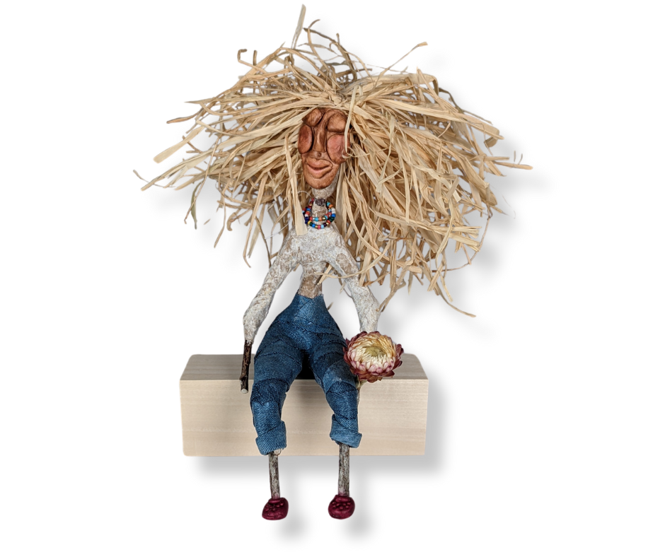 A whimsical spirit doll with a long paper mâché face, raffia hair, denim-style fabric pants, and tiny hot pink Crocs, sitting on a wood block holding a dried flower.
