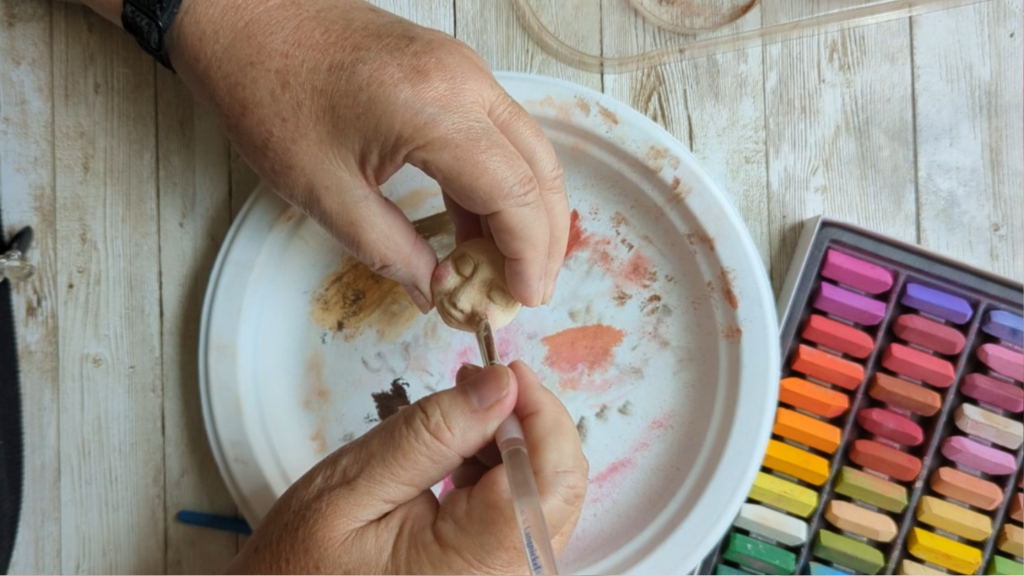 Hands painting the face of a handmade spirit doll face with soft pastels and a small brush. A process taught in The Artful Crone creative membership.
