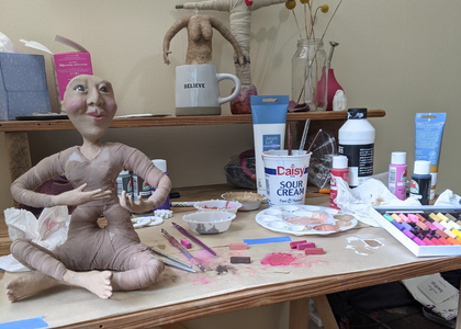 Spirit doll in progress on a worktable surrounded by clay, paint, and crafting materials.