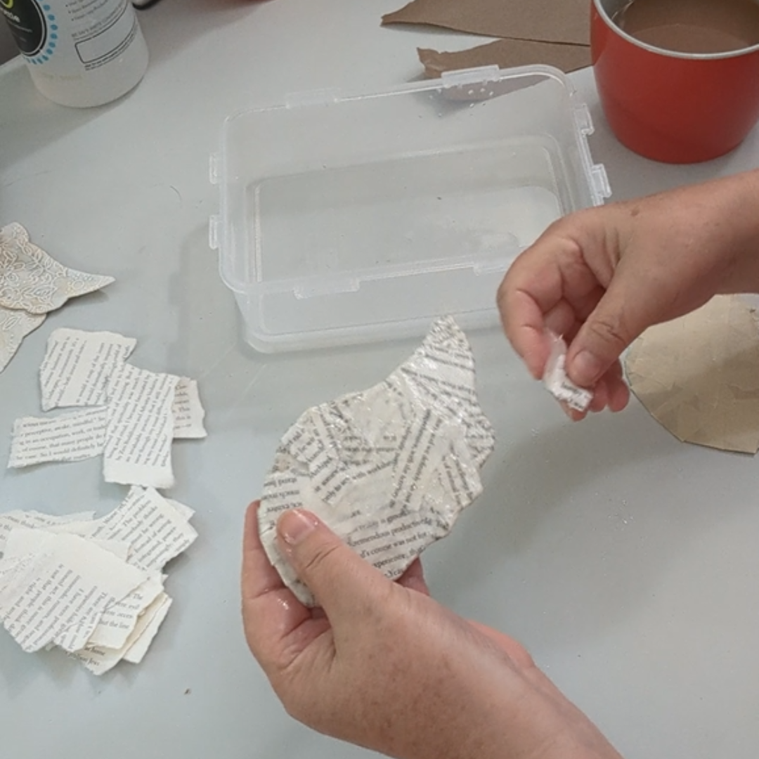 Hands adding torn book pages to a handmade angel wing during the papier-mâché process.