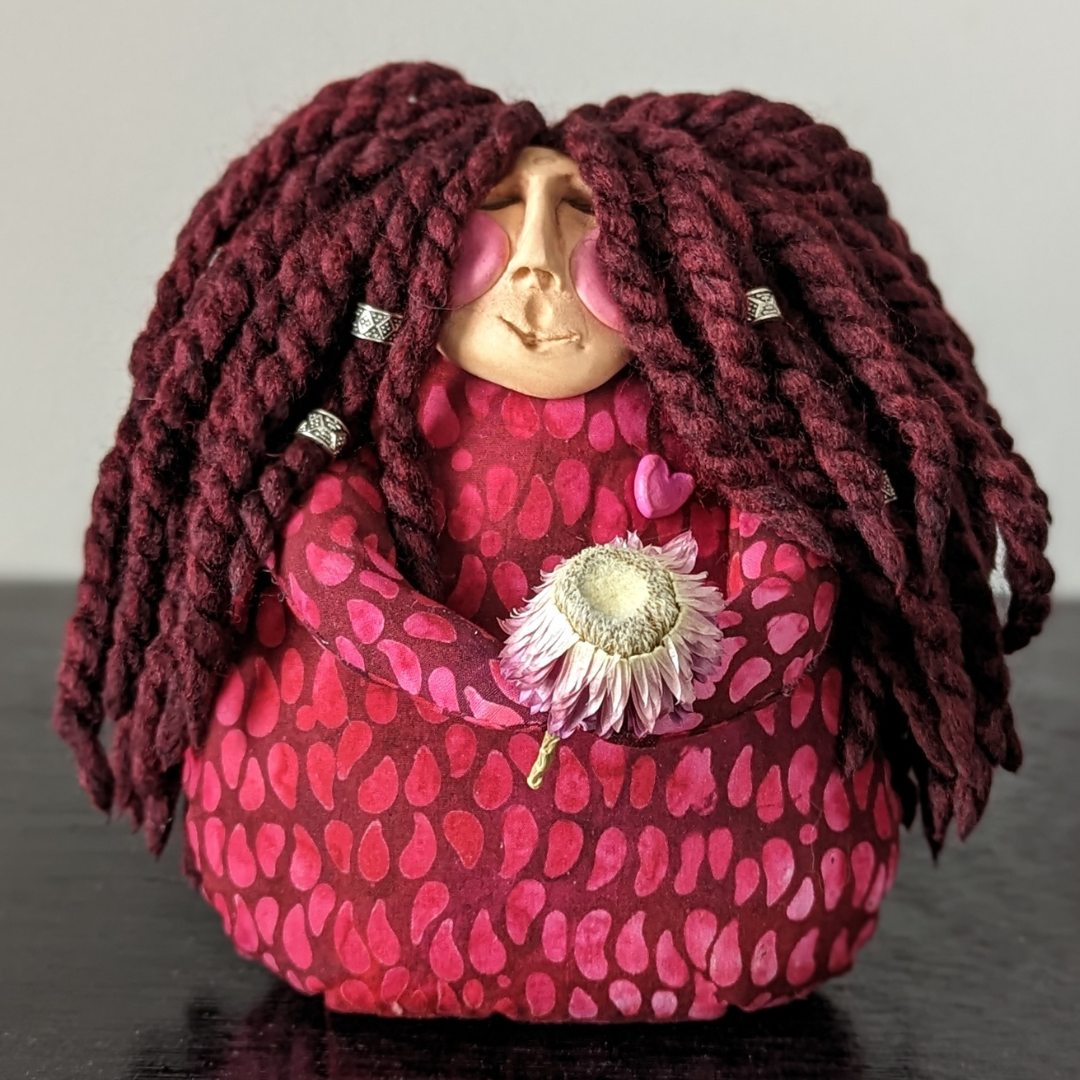 a small fabric doll with bright red fabric, a clay face and red yarn hair hoding a dried flower