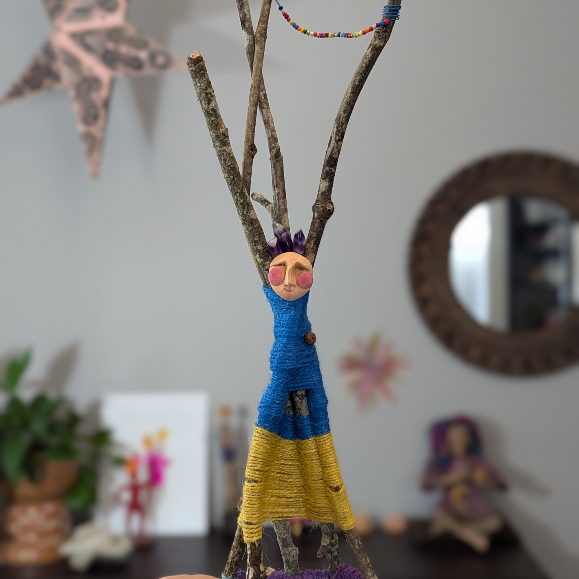 a spirit doll made from sticks wrapped with twine with a quirky clay face and crystals as a crown