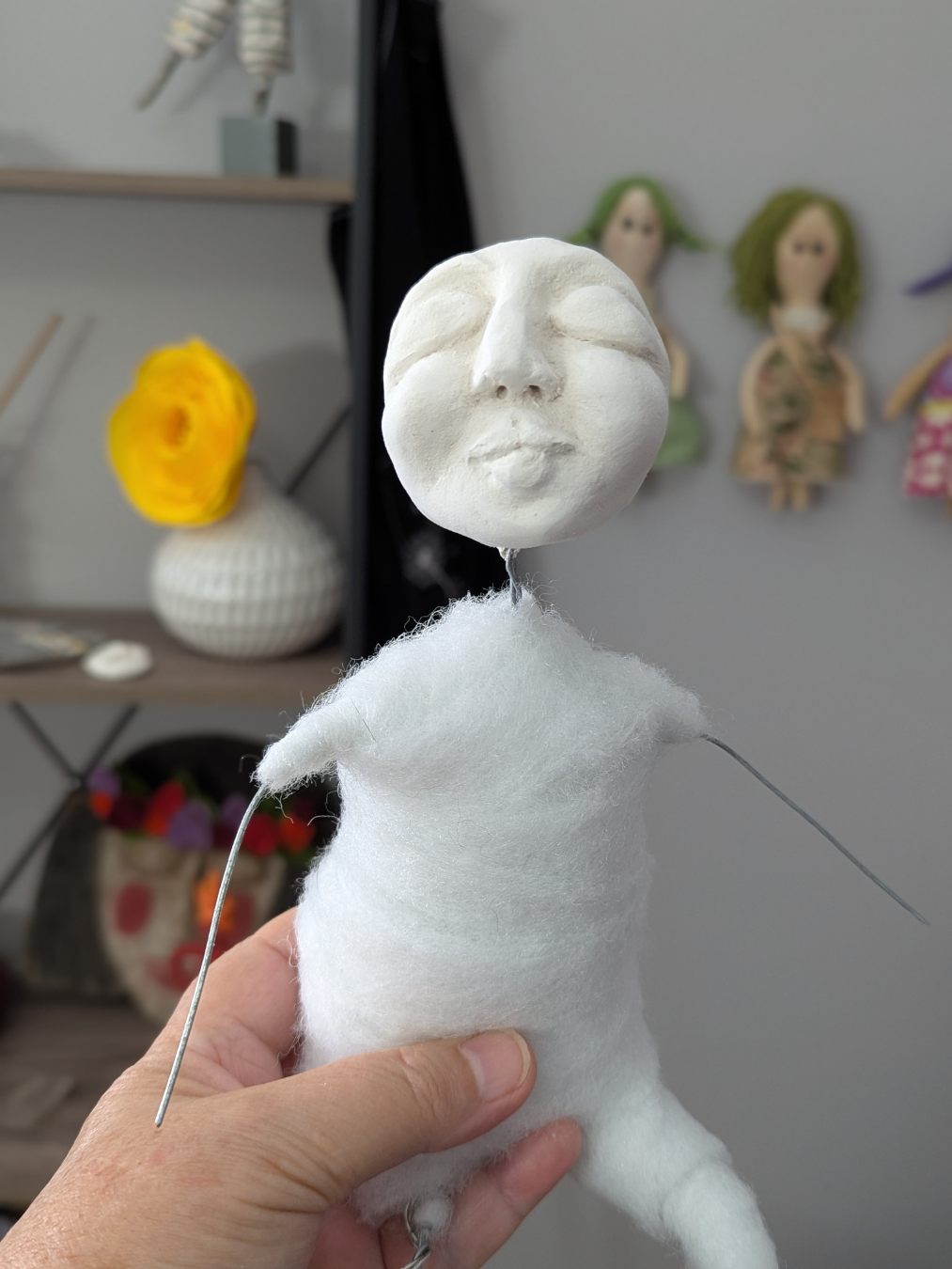 Handmade spirit doll in progress with a sculpted clay face, wrapped body, and exposed wire arms held in an artist’s hand.
