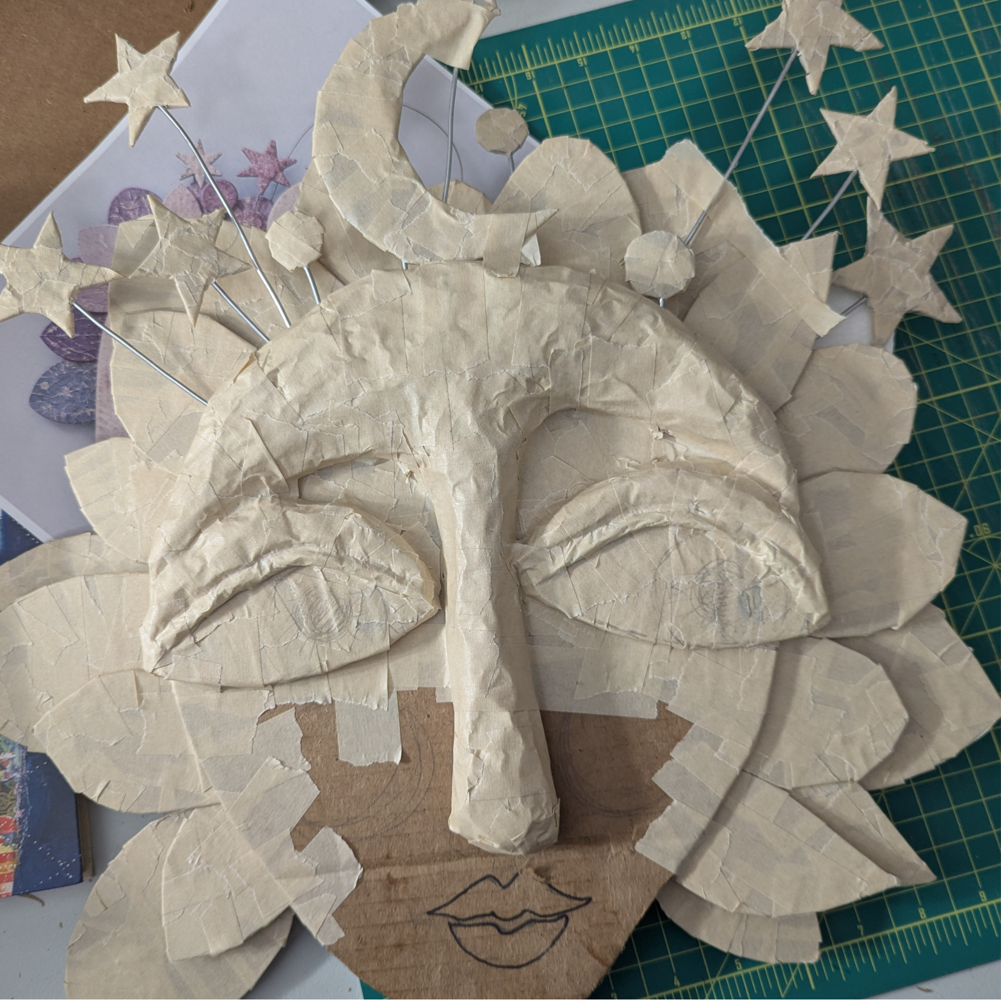 Paper mâché mask in progress with layered masking tape, closed eyes, a long nose, moon and star shapes, and wire details on a worktable.