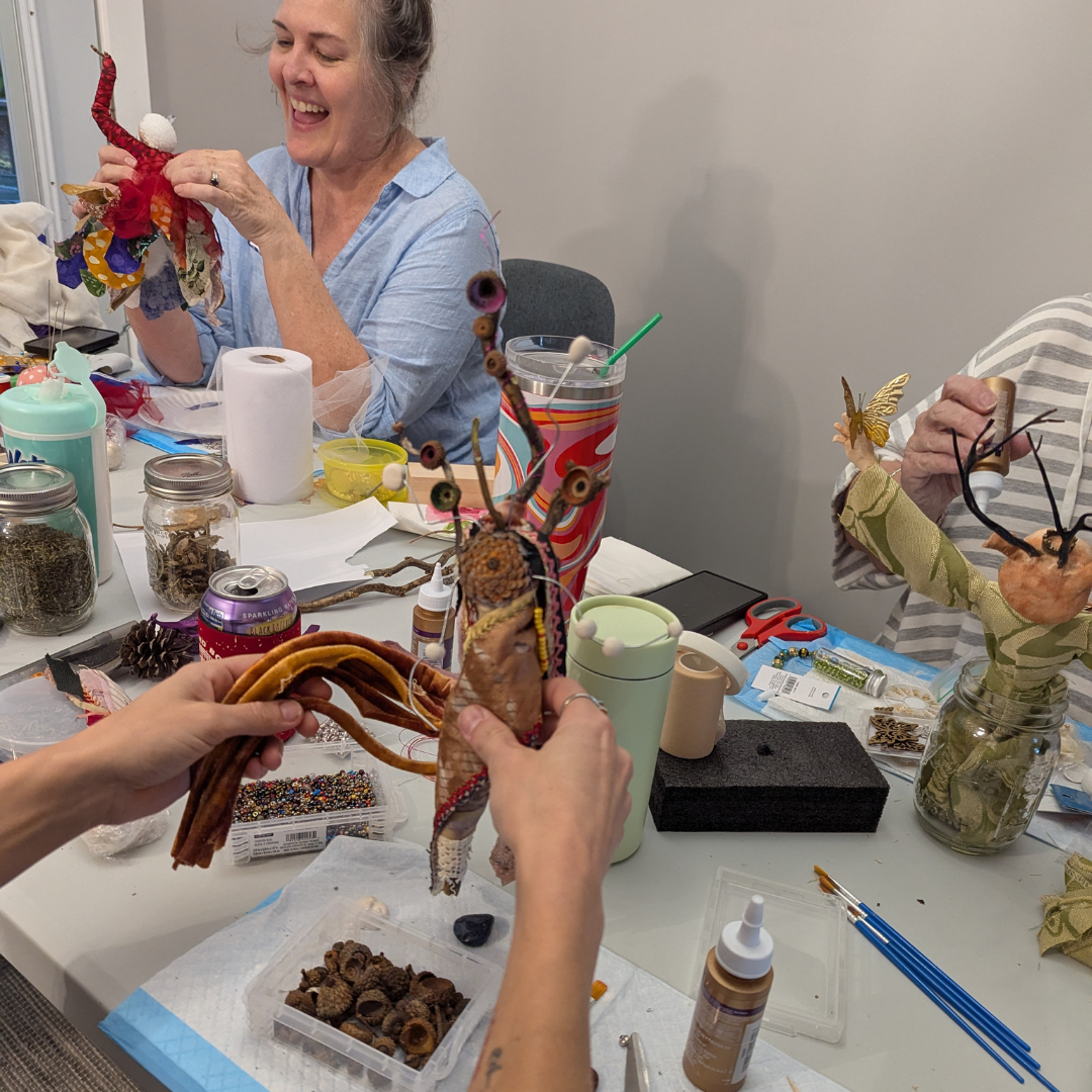 Women making stick doll spirit dolls with a third holding up her doll. The doll's arms are stretched out and the face is round with ears and a green robe