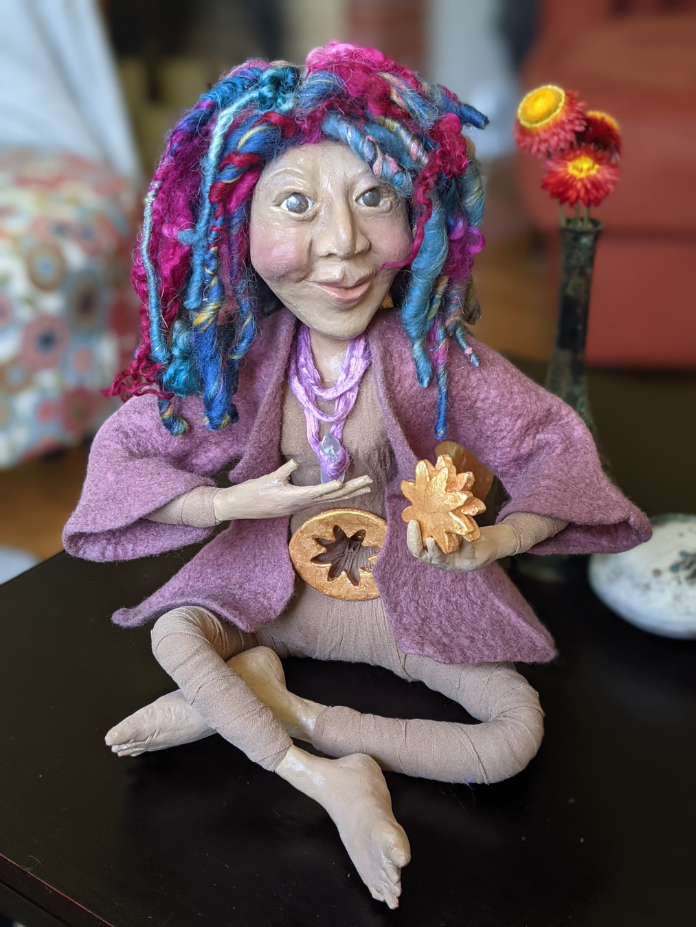 Handmade seated spirit doll with colorful yarn hair, a pink cloak, and small golden symbols in her hands.