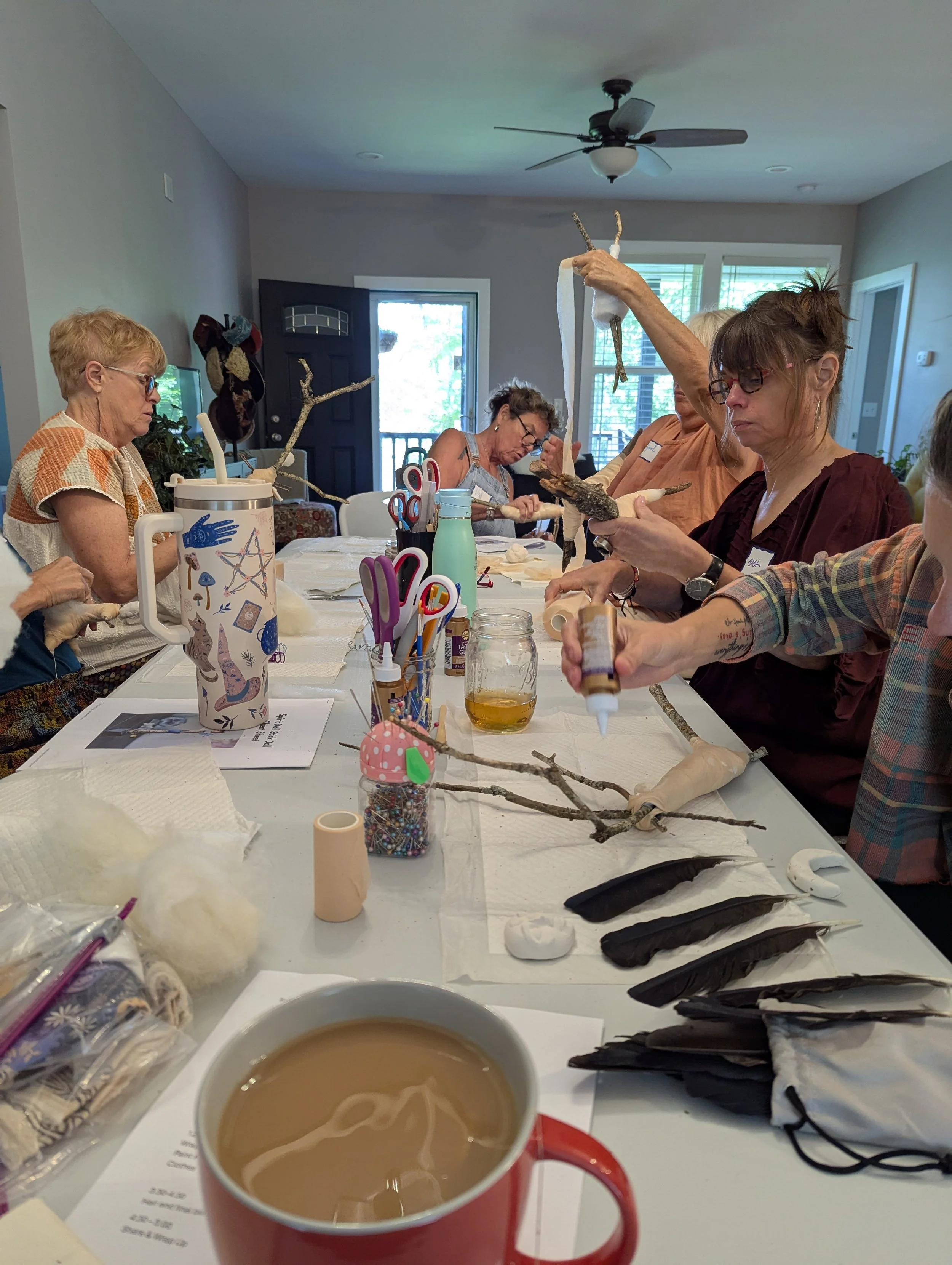 Close-up view of participants at a spirit doll workshop, wrapping sticks and shaping fabric in a light-filled studio. Creative supplies and sculpted clay faces are spread across the table.