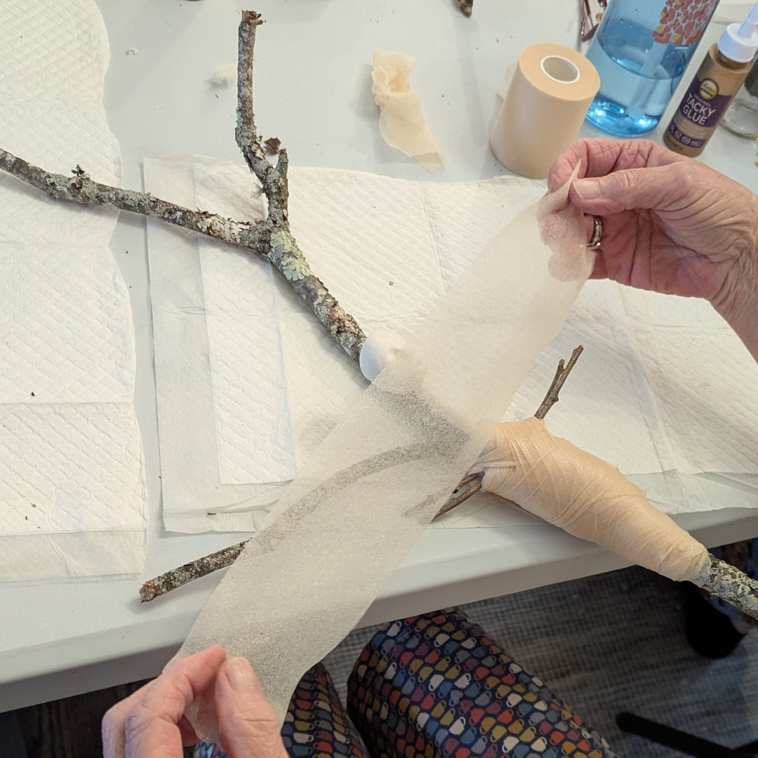 Hands wrapping batting around a stick doll body to build shape before adding fabric and clothing.