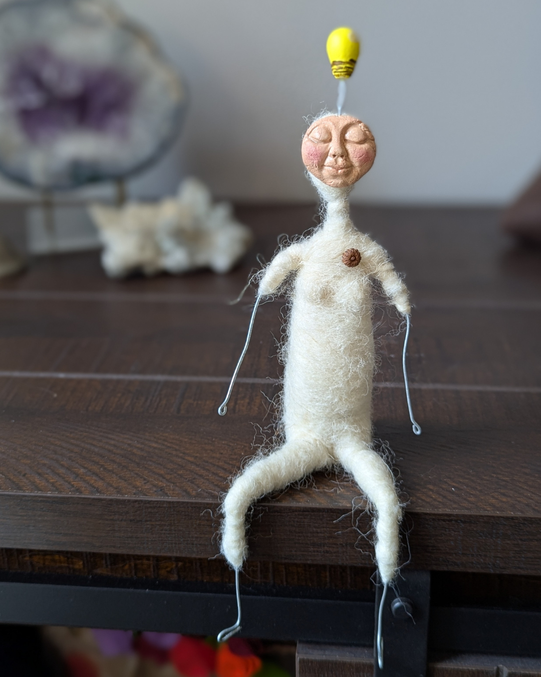 A small needle felted spirit doll sitting on a table with a clay lightbulb emerging from the top of her head, symbolizing inspiration and insight.