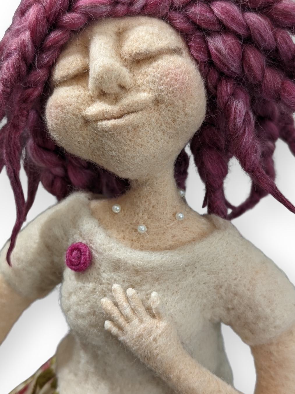 A close-up of a handmade spirit doll with braided magenta wool hair, a serene smiling expression, pearl-like beaded necklace, and one hand placed over her heart. Her soft felted texture and peaceful face convey tenderness and intuitive wisdom.