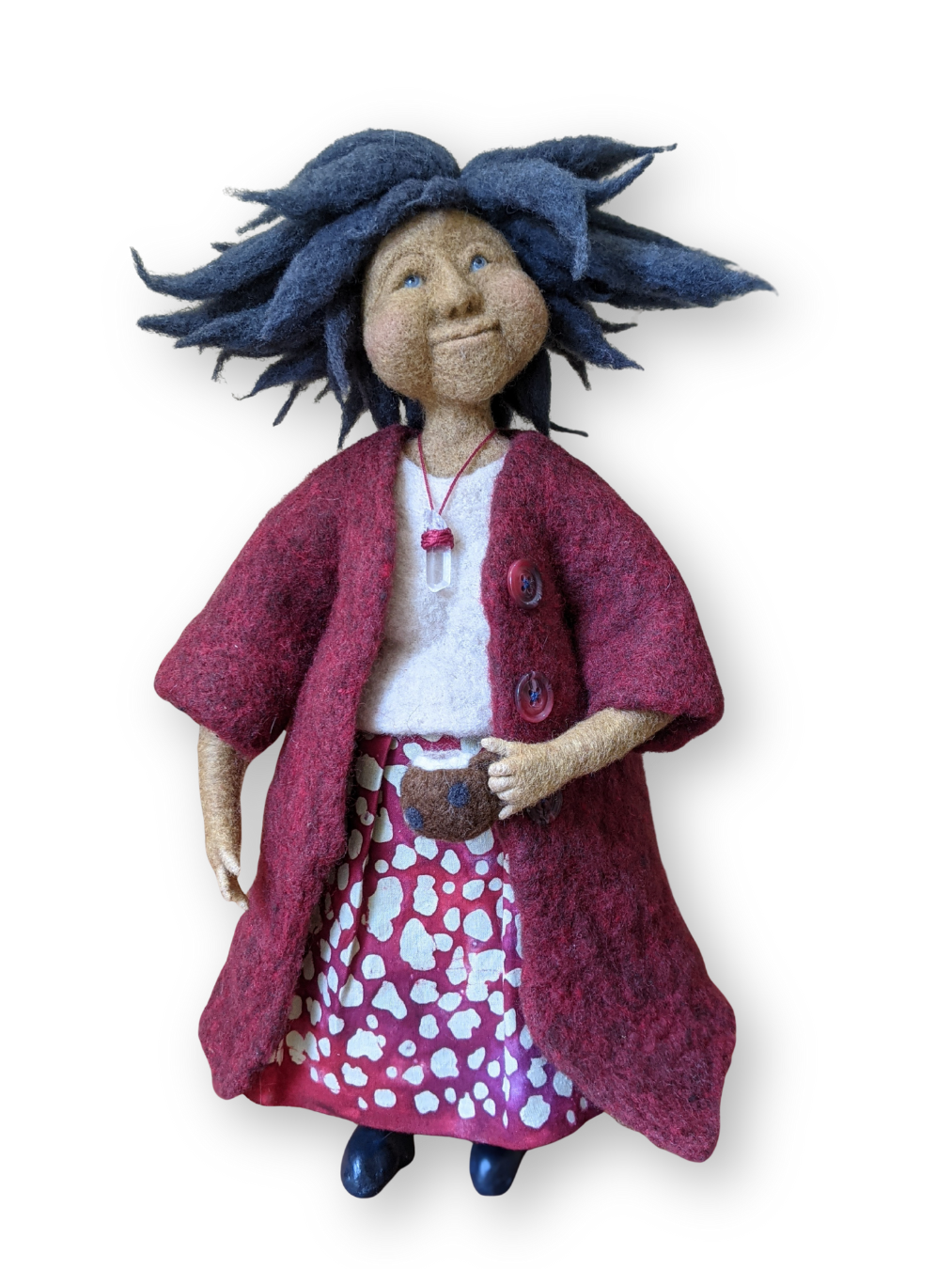 A handmade needle-felted spirit doll with tousled blue hair, a soft red felt coat, and a patterned red skirt, holding a tiny felt mug. She stands with a gentle, knowing smile, embodying warmth, groundedness, and soulful presence.