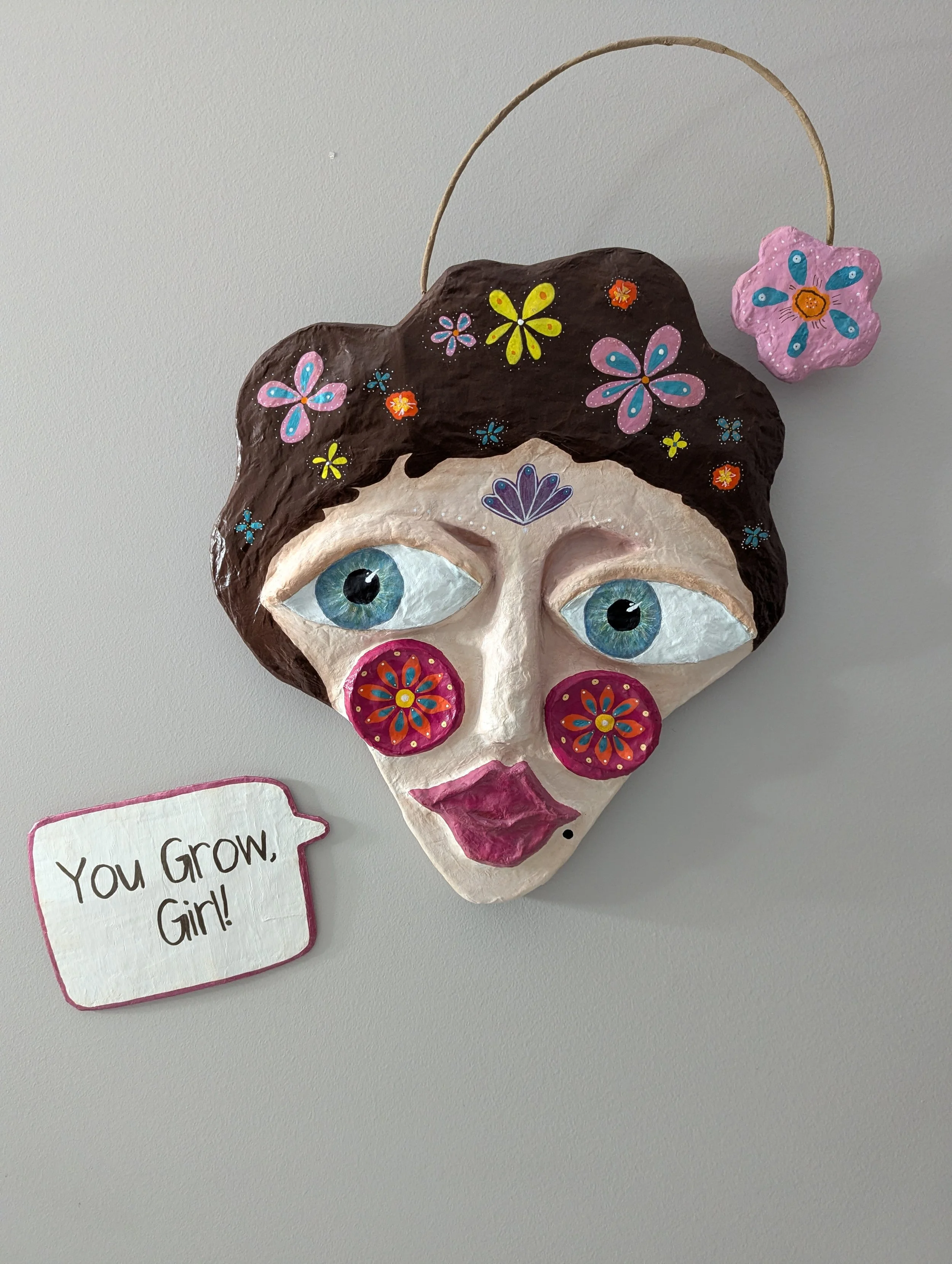 Colorful handmade papier-mâché spirit mask with painted flowers, blue eyes, pink cheeks, and a small sign that says You grow, girl.