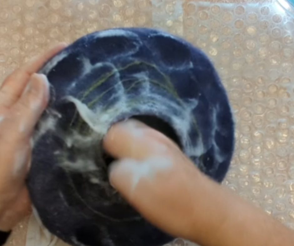 Hands shaping a dark blue wet-felted bowl, pressing into the center to form the vessel