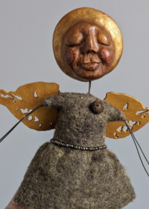 Handmade spirit doll with a round golden face, closed eyes, and a peaceful expression, mounted above a felted wool body with small wings and simple wire arms.