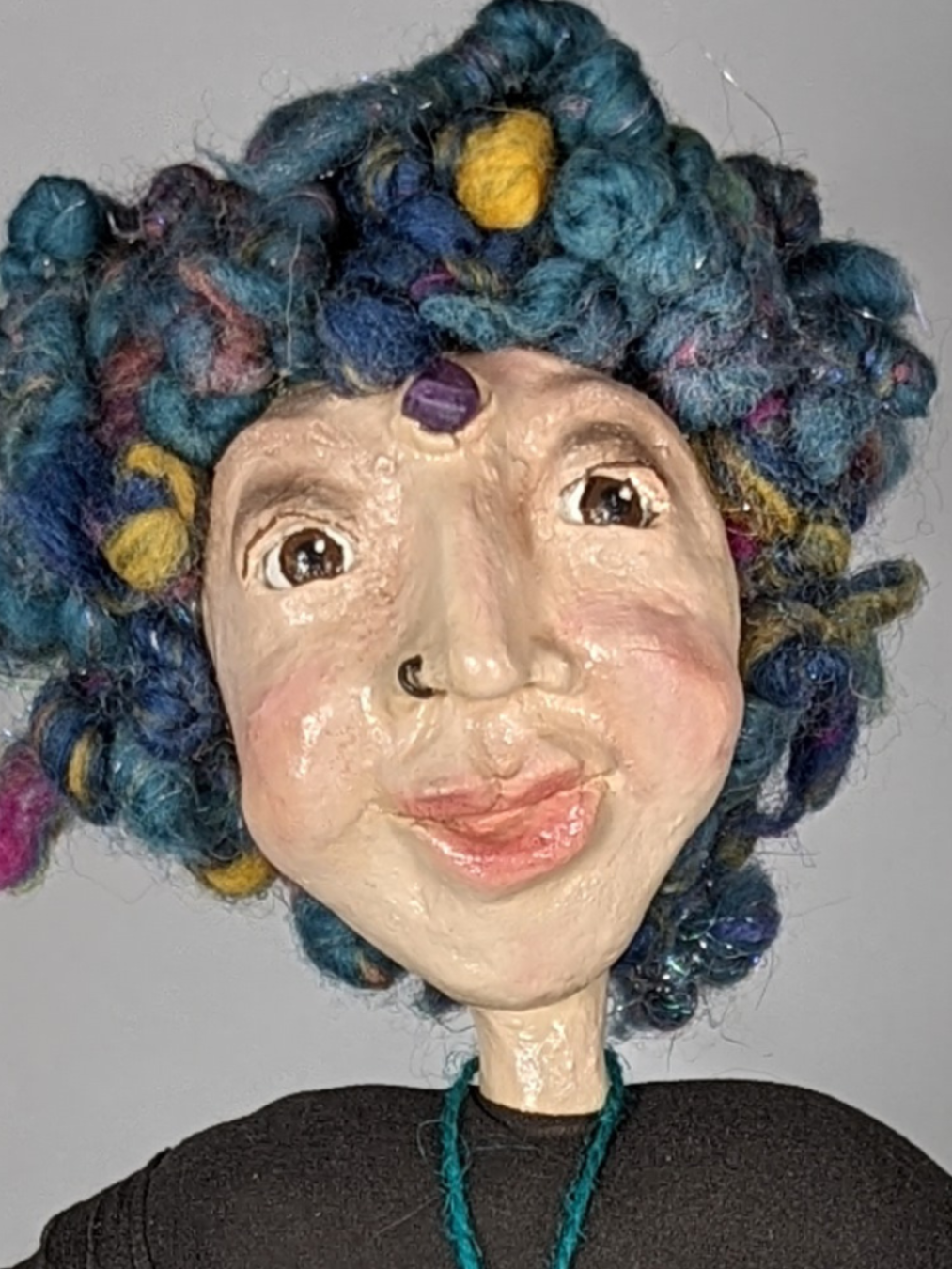 a close up of a spirit doll made of clay. The doll has a nosering, a purple bead on her third eye and colorful blue hair. Made by Stacy Vajta