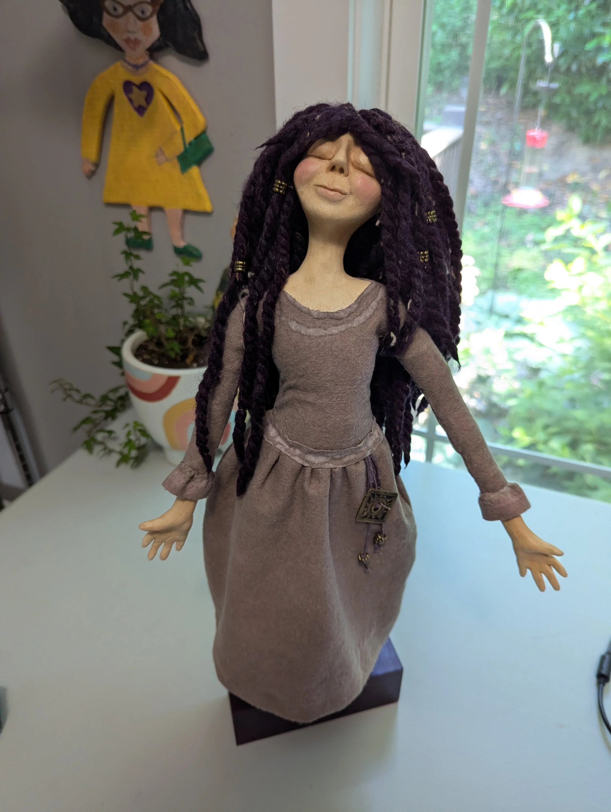 A finished handmade spirit doll with a soft fabric dress, standing near a studio window.