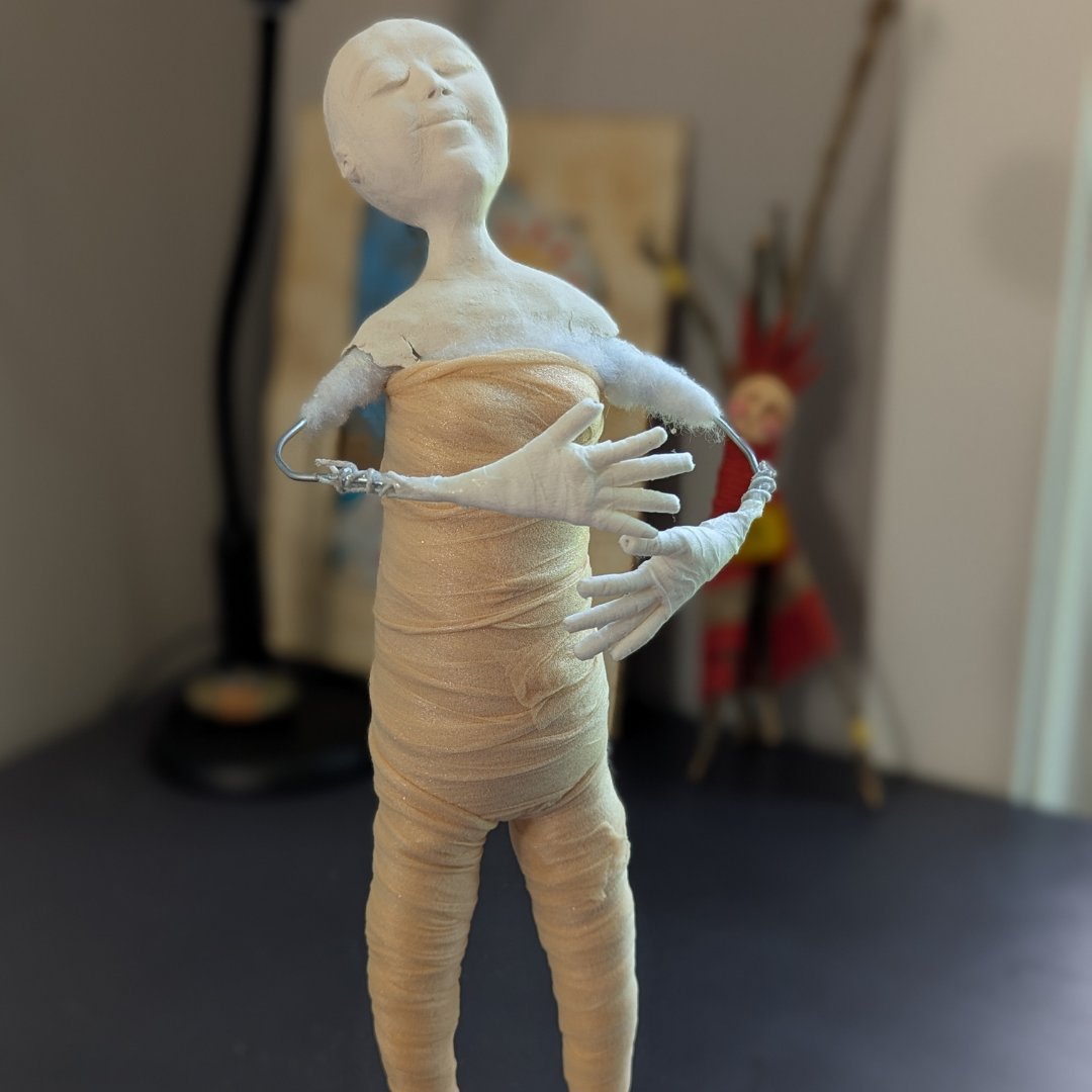Spirit doll in progress with a sculpted clay head and hands, wrapped fabric body, and partially exposed wire arms.