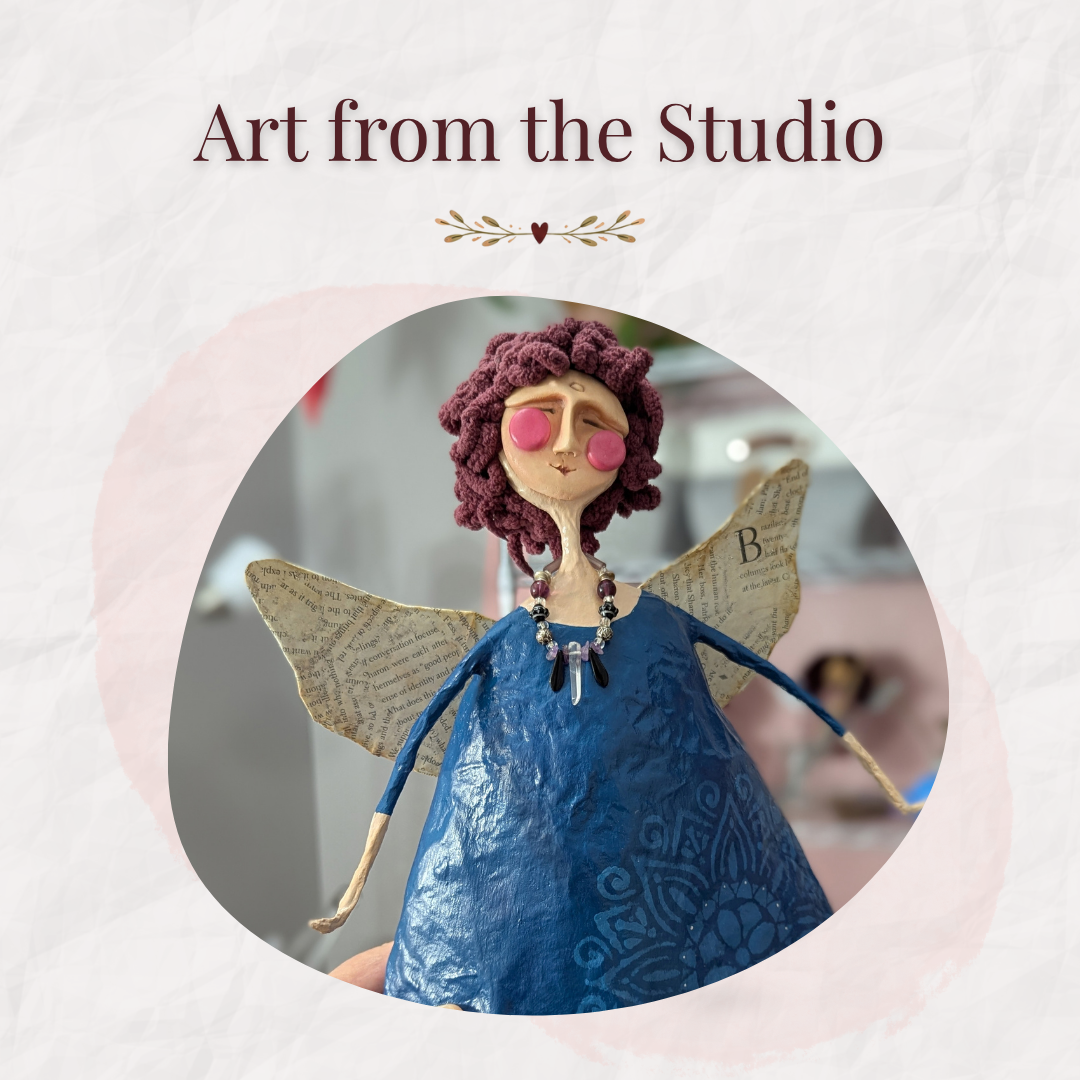 Handmade papier-mâché angel with a deep blue dress, burgundy yarn hair, paper wings made from printed book pages, and a beaded necklace, beneath the words “Art from the Studio.”