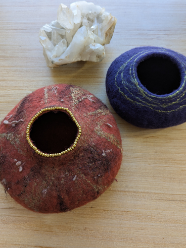 Handmade felted wool intention vessels in earthy red and deep blue with stitched details, resting on a wooden surface beside a clear quartz crystal.