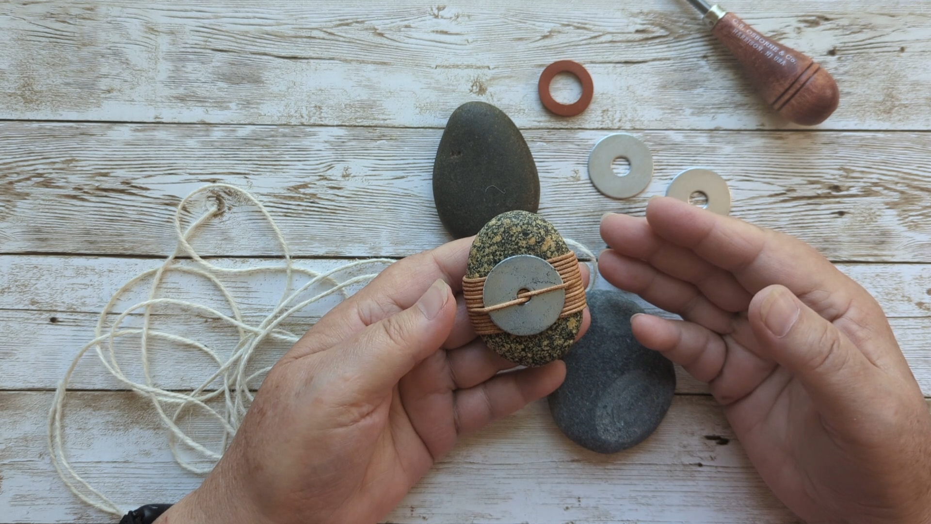 Hands holding a small stone talisman wrapped with copper wire and a metal disc, surrounded by natural stones and simple tools on a wooden surface.