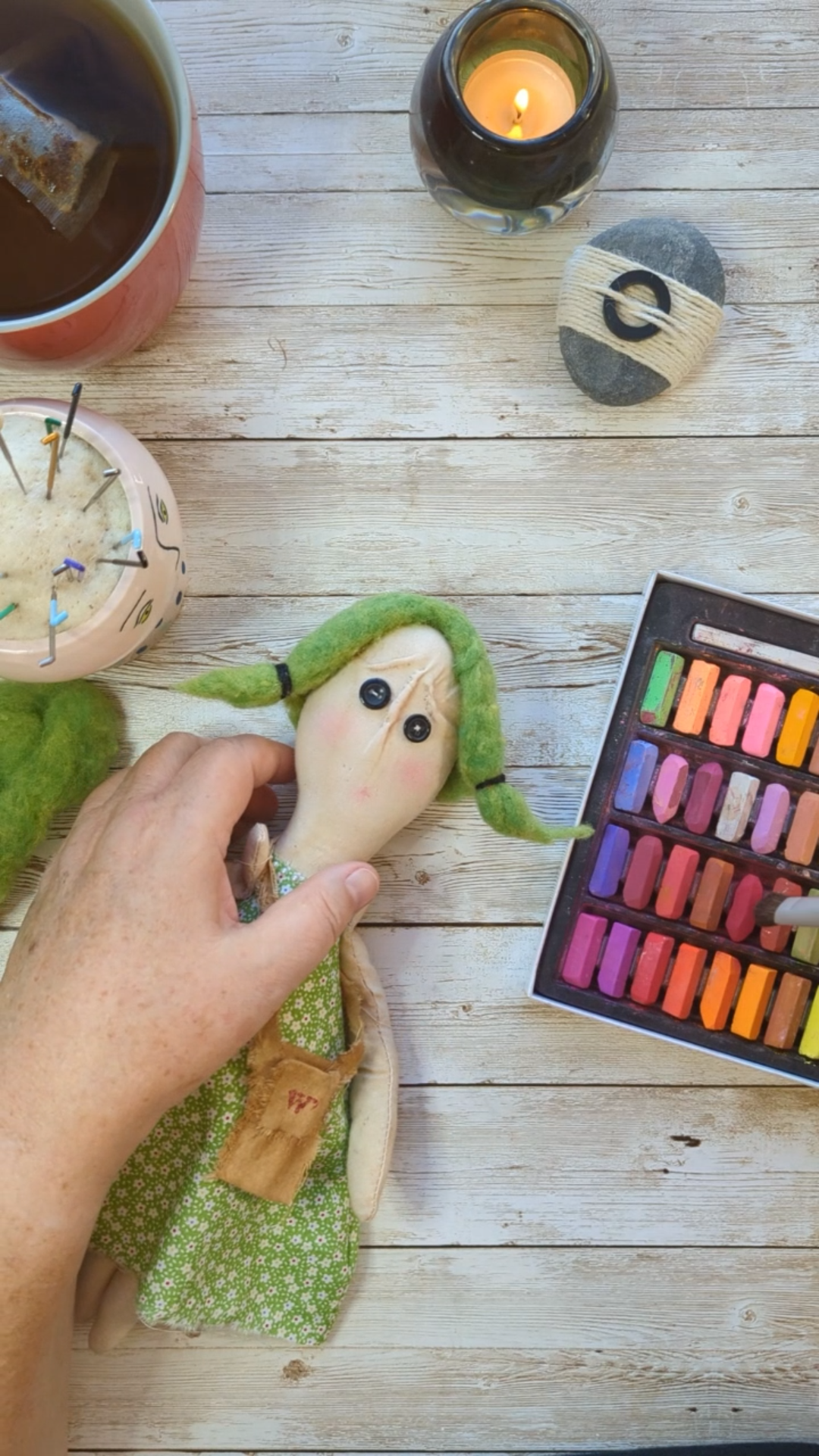 Hands coloring the face of a handmade soft doll using chalk pastels, with tea, candle, and creative tools nearby.