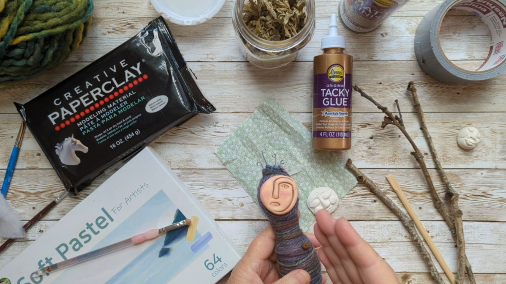Hands holding a small wrapped spirit doll in progress, surrounded by creative materials including paperclay, soft pastels, tacky glue, dried herbs, twigs, and tools on a wooden tabletop.