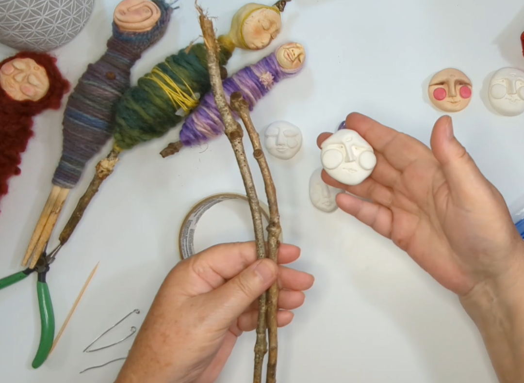 Small clay face held beside two sticks, with several finished Bundle Dolls and face options on the worktable.