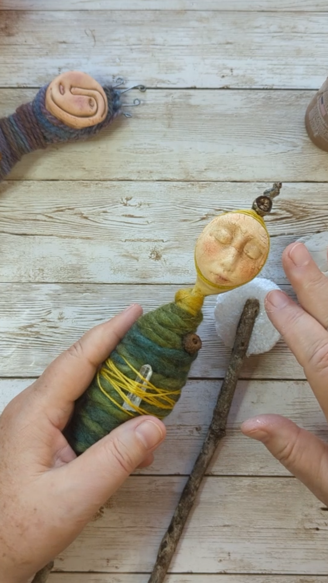 Hands holding a wool-wrapped spirit doll with a sculpted face, crystal, and twig elements during a talisman-making process.