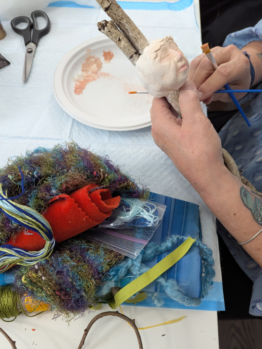 Hands painting a clay face on a stick doll during a mixed-media spirit doll workshop, with yarn, fabric, scissors, and art supplies on the table.