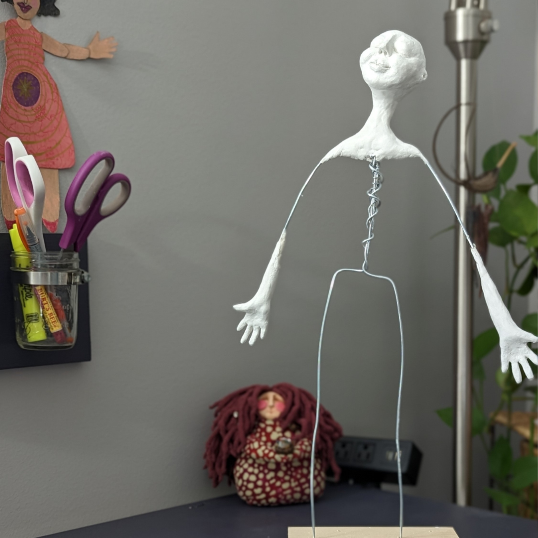 Spirit doll in progress with a wire armature, sculpted clay head, and clay hands standing on a wooden base in an artist’s studio.