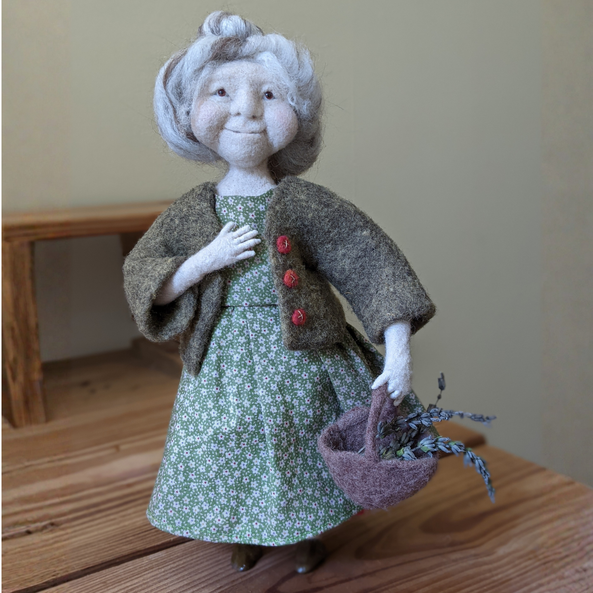 a needle felted doll of a gransmother holding a basket of lavender wearing a green dress with green sweater and her haor in a bun standing on a wood table