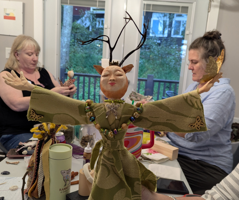 Two women making stick doll spirit dolls with a third holding up her doll. The doll's arms are stretched out and the face is round with ears and a green robe
