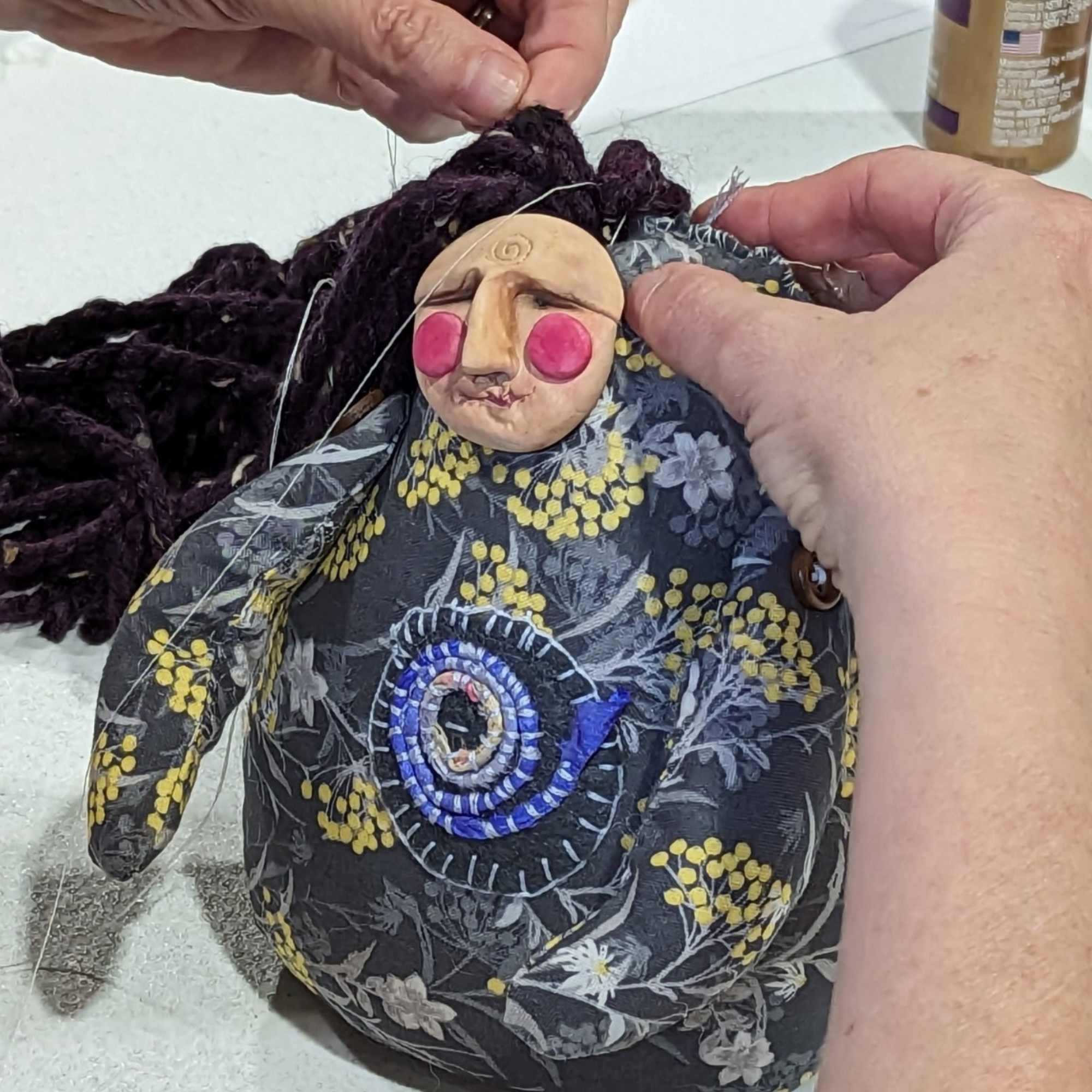 Close-up of hands stitching yarn hair onto a handmade poppet doll during a live workshop.