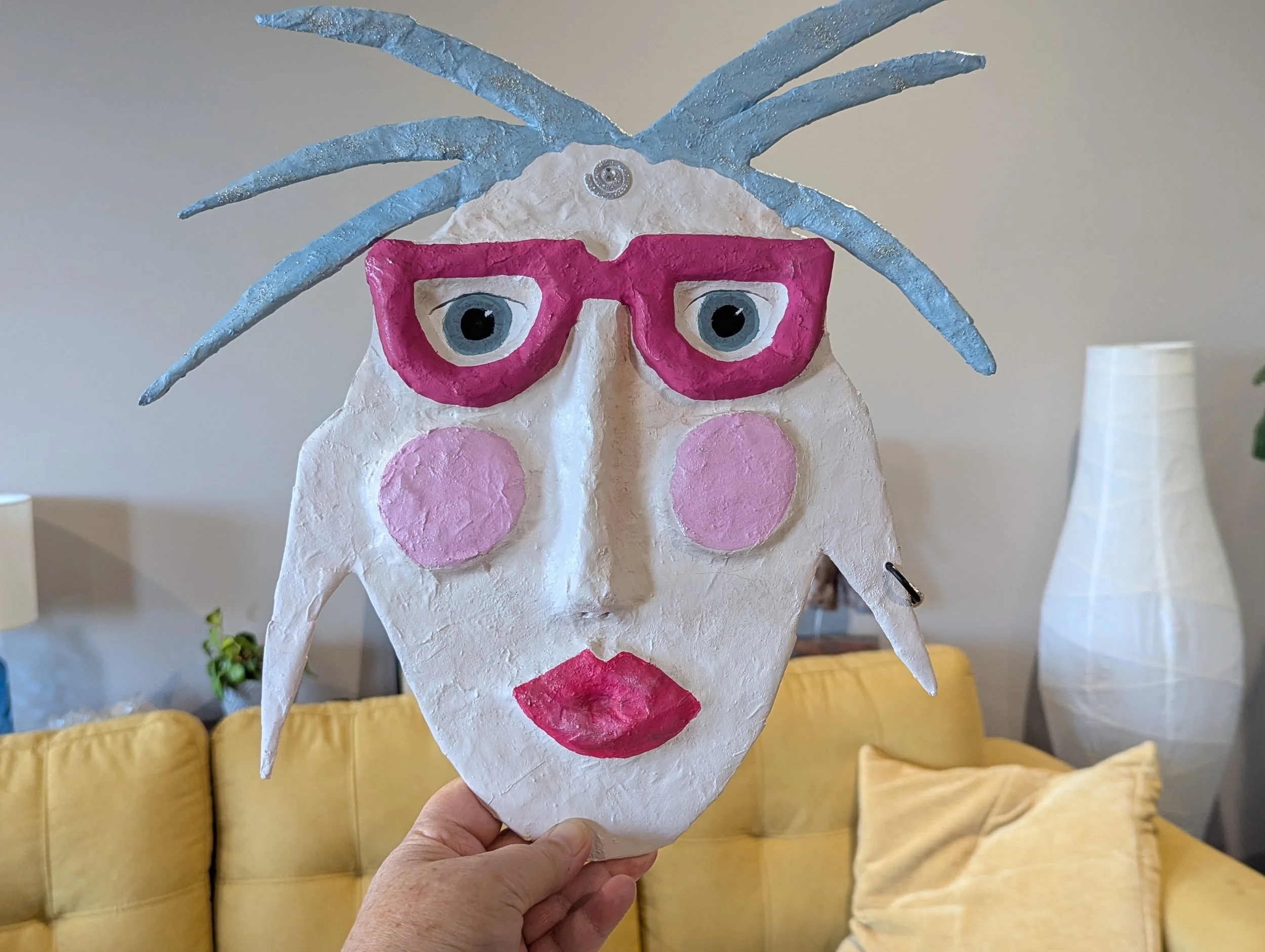 A hand holding a whimsical paper mache mask with bright pink glasses, blue spiky hair, rosy cheeks, and bold pink lips, against a cozy living room background.