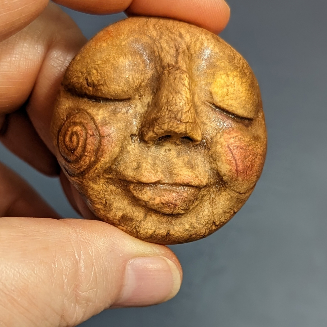 Finished painted clay face with closed eyes, rosy cheeks, and a spiral detail.