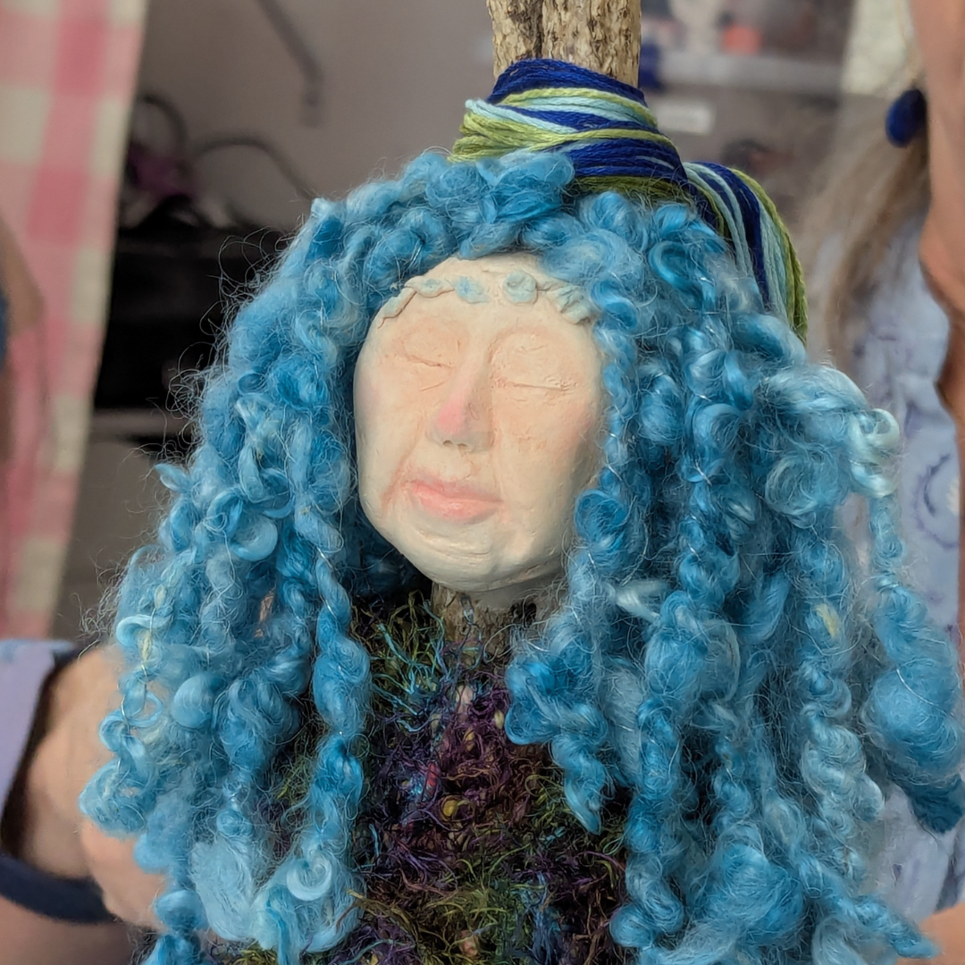 Finished stick doll with a soft clay face, bright blue curly yarn hair, wrapped fibers, and colorful textile details.