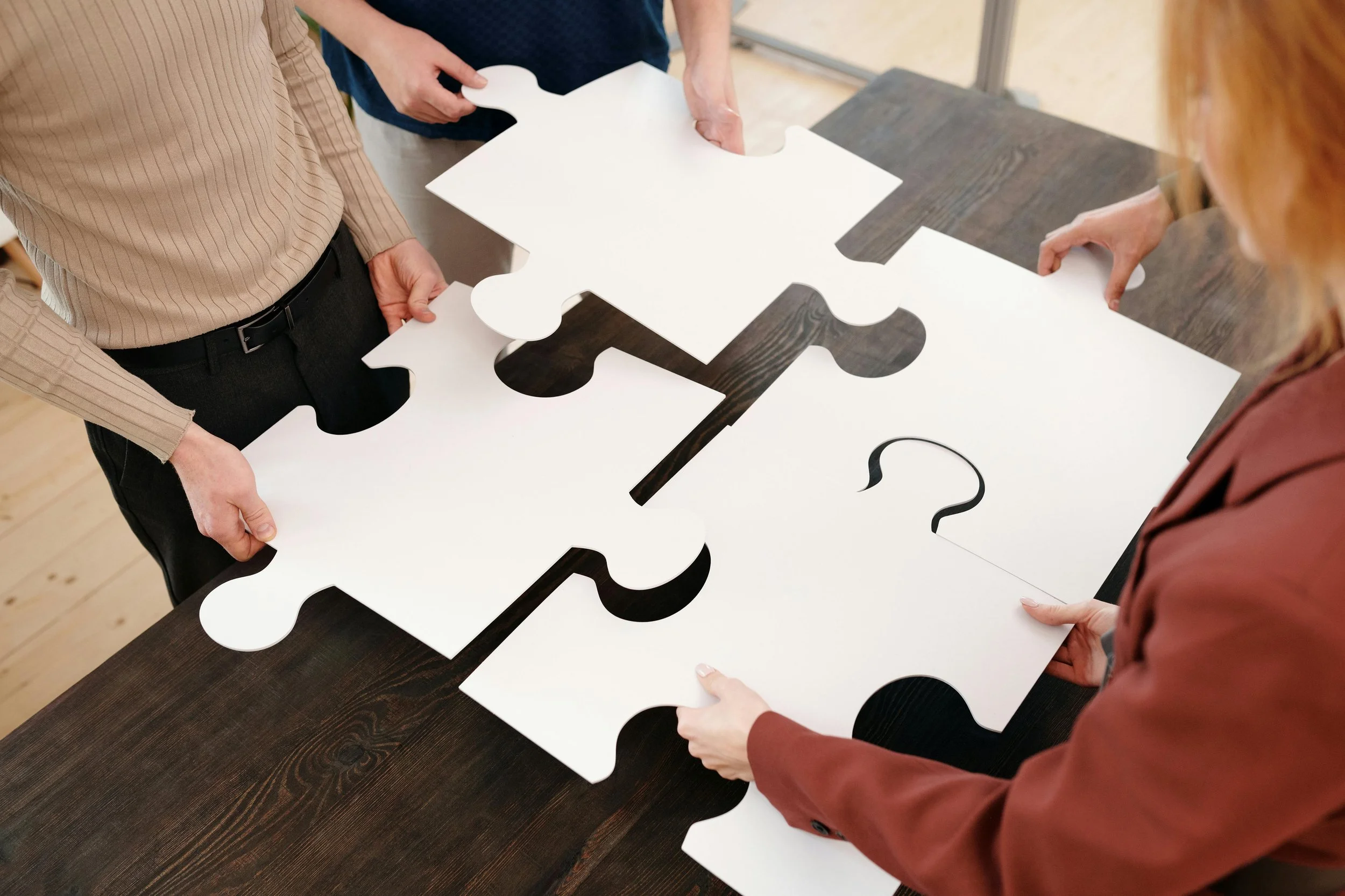 Four people assembling four large white jigsaw puzzle pieces on a dark wooden table. Representing healing, connection, therapy, personal growth.
