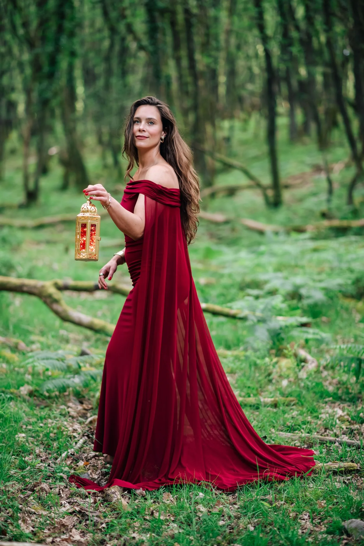 A woman in a flowing red gown standing in a green forest, holding a decorative lantern.