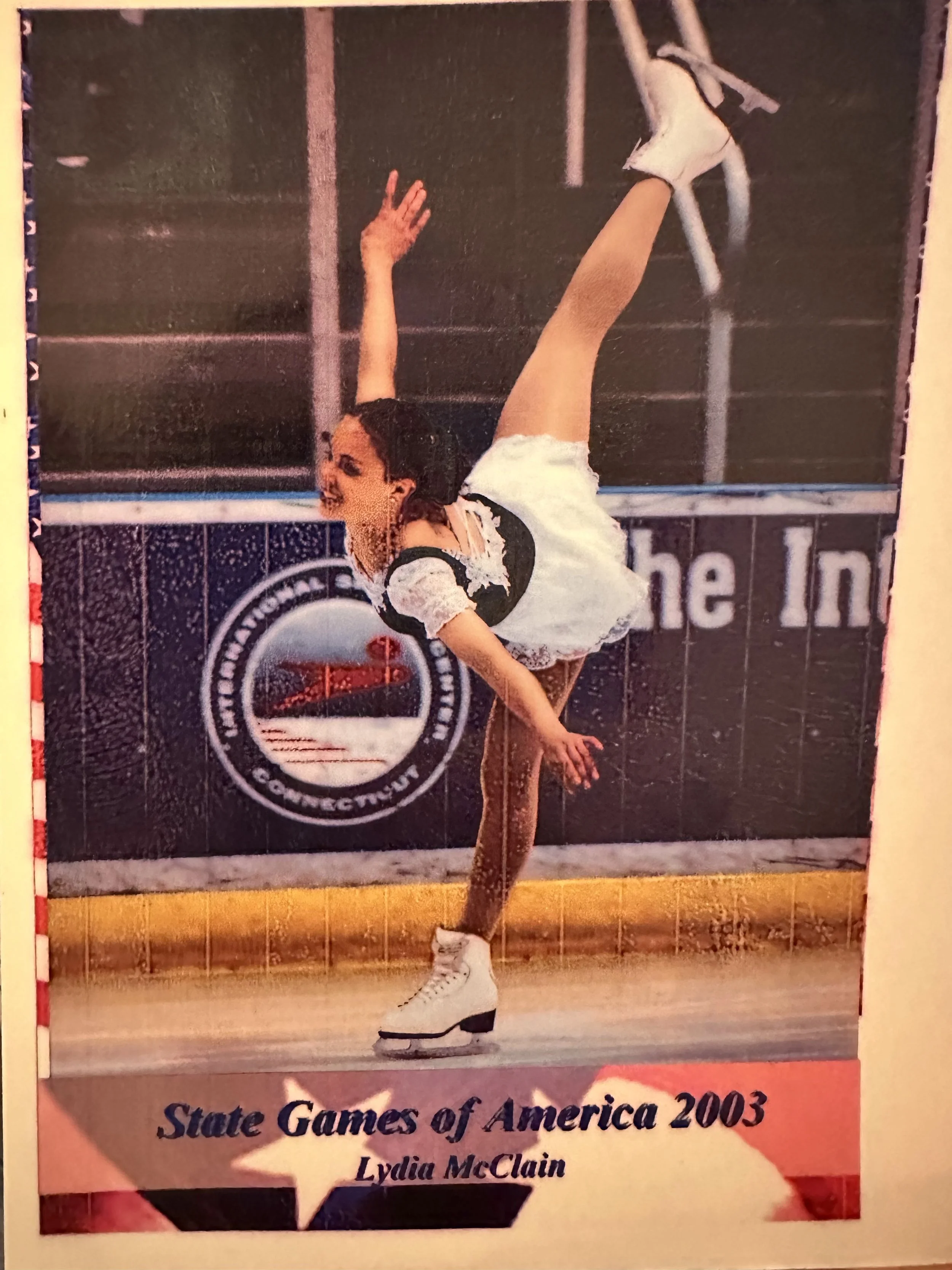 A young woman figure skater on ice in a pose, wearing a black and white costume and white ice skates, with her right arm raised and left arm extended downward, standing on one leg with the other leg raised behind her during a performance at the 2003 State Games of America.