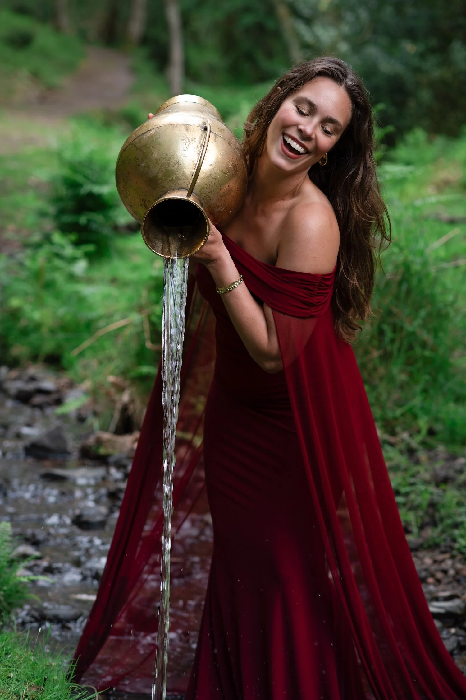 A woman in a red dress standing outdoors, smiling, and pouring water from an old brass teapot.