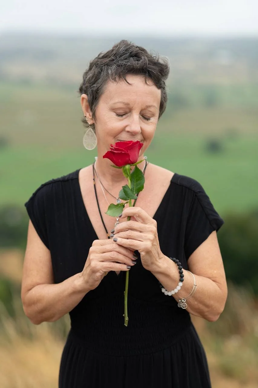 A woman with short gray hair holding a red rose close to her face with her eyes closed, standing outdoors with a blurred green landscape in the background.