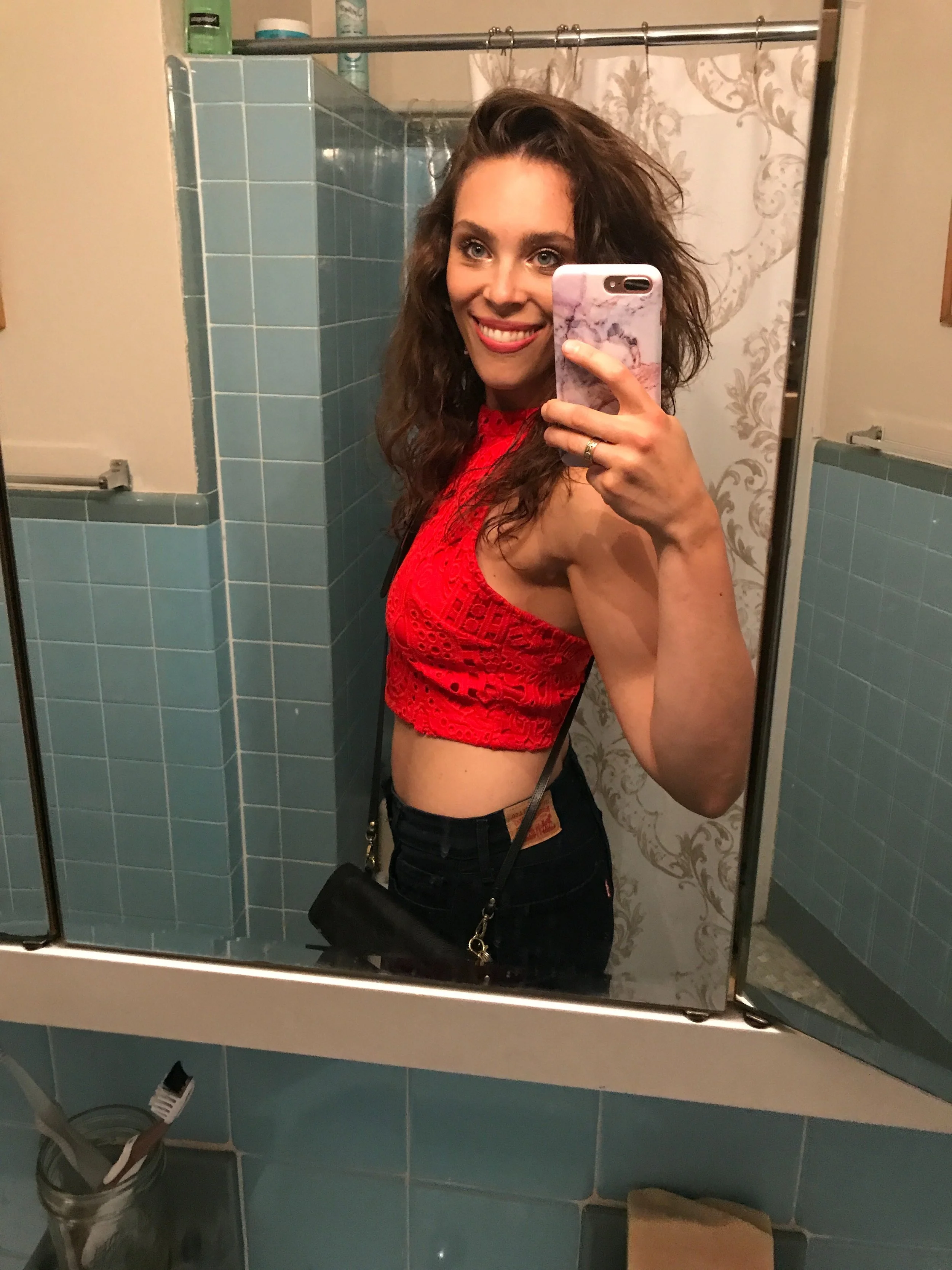 A woman taking a mirror selfie in a bathroom, wearing a red top and black pants, with curly hair and holding a phone with a pink marble case. Bathroom features include teal tiles, a shower with a curtain, and toiletries on the shelf.