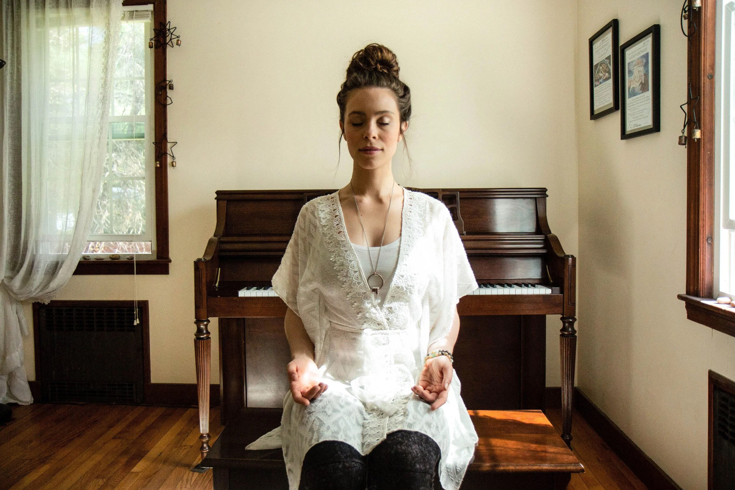 A young woman with dark hair styled in a top knot, sitting with eyes closed and hands resting on her lap, wearing a white lace dress and a pendant necklace, positioned in front of a vintage upright piano inside a room with wooden floors, a window with sheer curtains, and framed artwork on the wall.