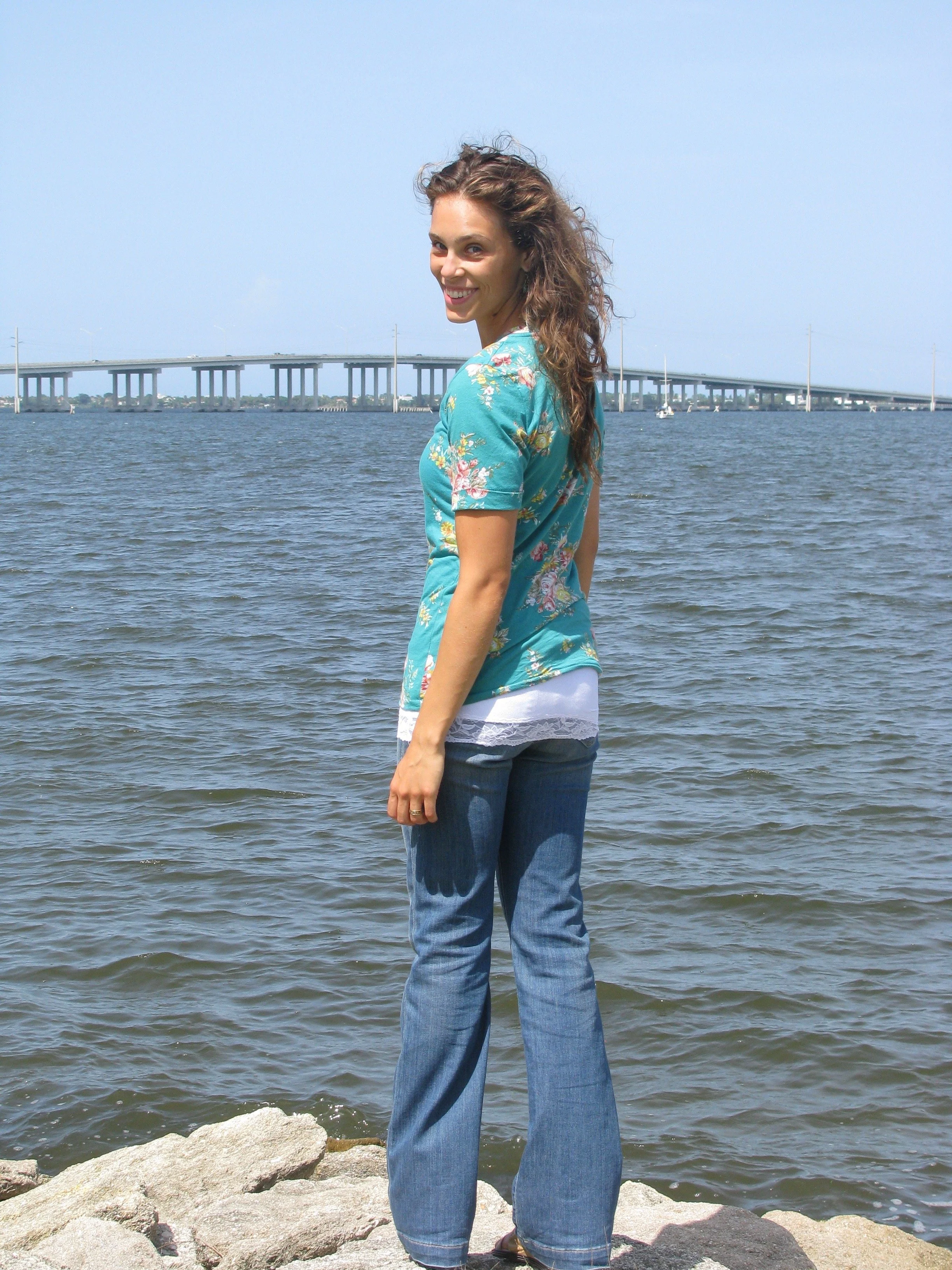 A woman with curly brown hair smiling, standing on rocks by a body of water with a bridge in the background.